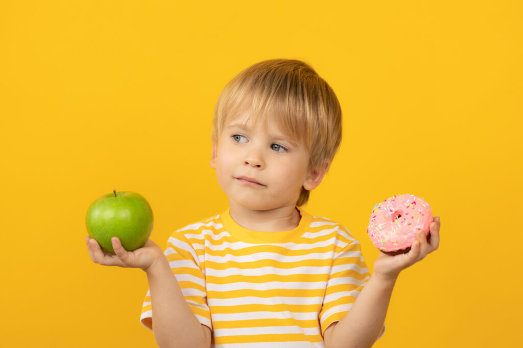 Happy child holding donut and apple. Portrait of funny kid against yellow background. Healthy food concept. Difficult choice